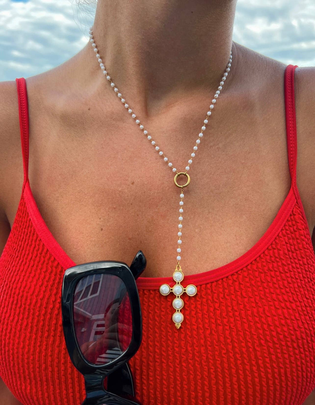 Person wearing a red tank top and pearl necklace with a cross pendant, holding sunglasses.