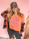 Woman wearing an orange graphic t-shirt with text, black cap, and plaid jacket against a pink background