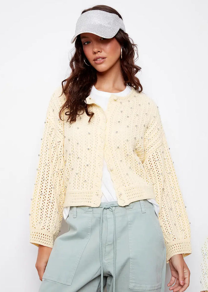 Woman wearing a light yellow crochet cardigan over a white shirt with light blue pants on a white background.