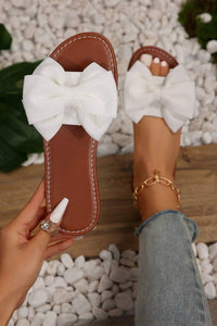 Brown sandals with large white bows held by a hand on a pebbled surface.