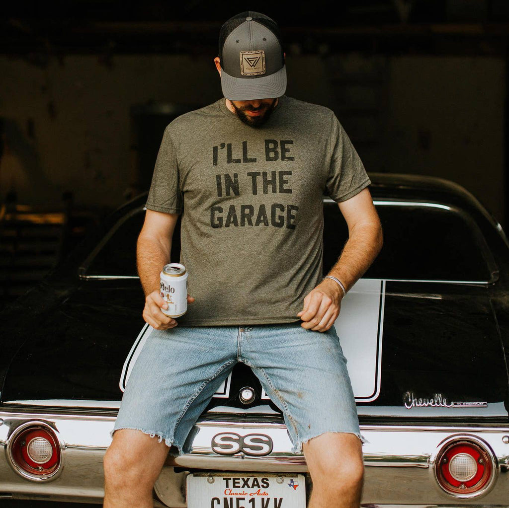 Man wearing a t-shirt with 'I'll be in the garage' text, standing next to a classic car.
