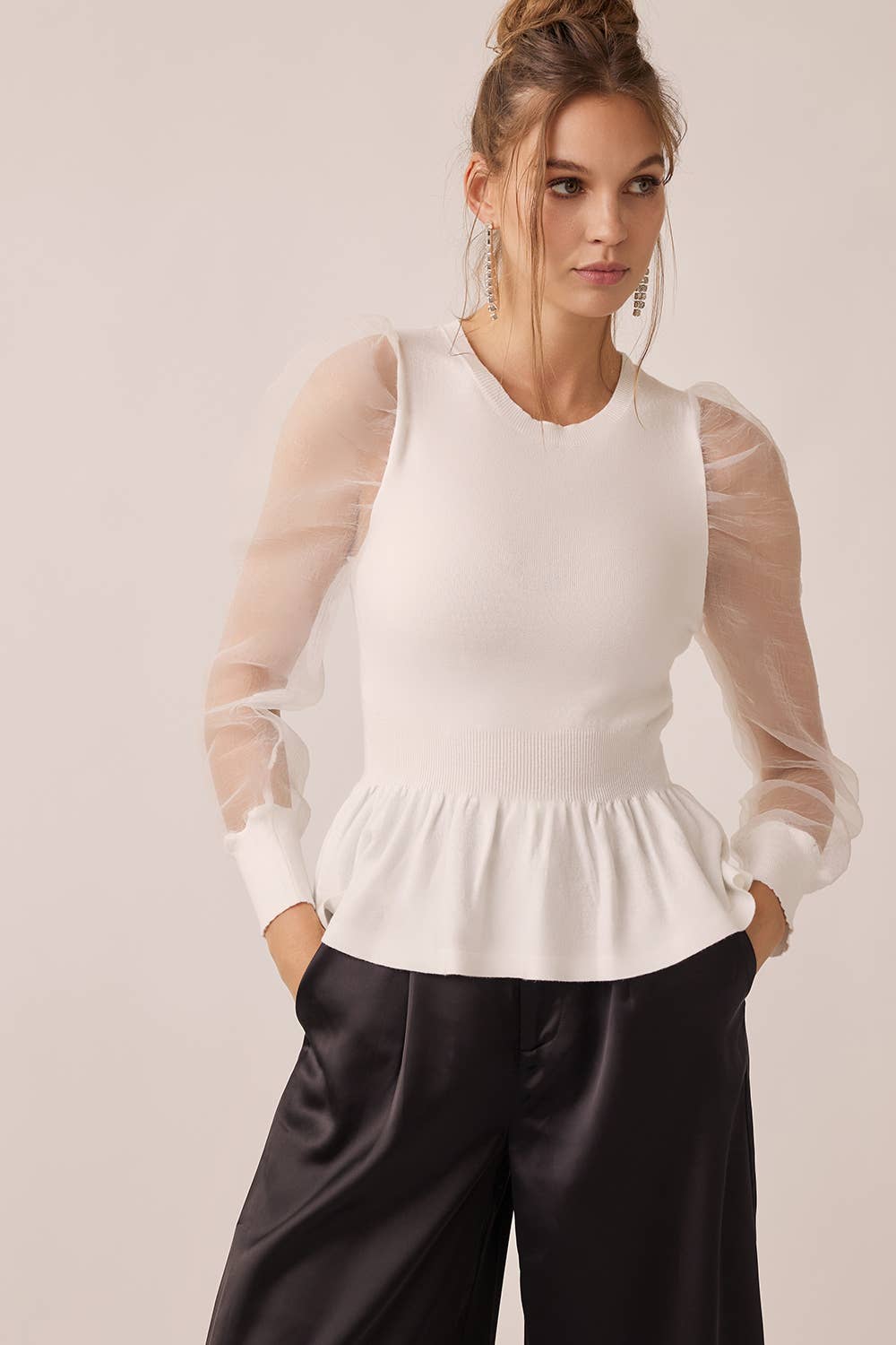 Woman wearing a white top with sheer sleeves and black pants on a light background