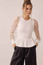Woman wearing a white top with sheer sleeves and black pants on a light background