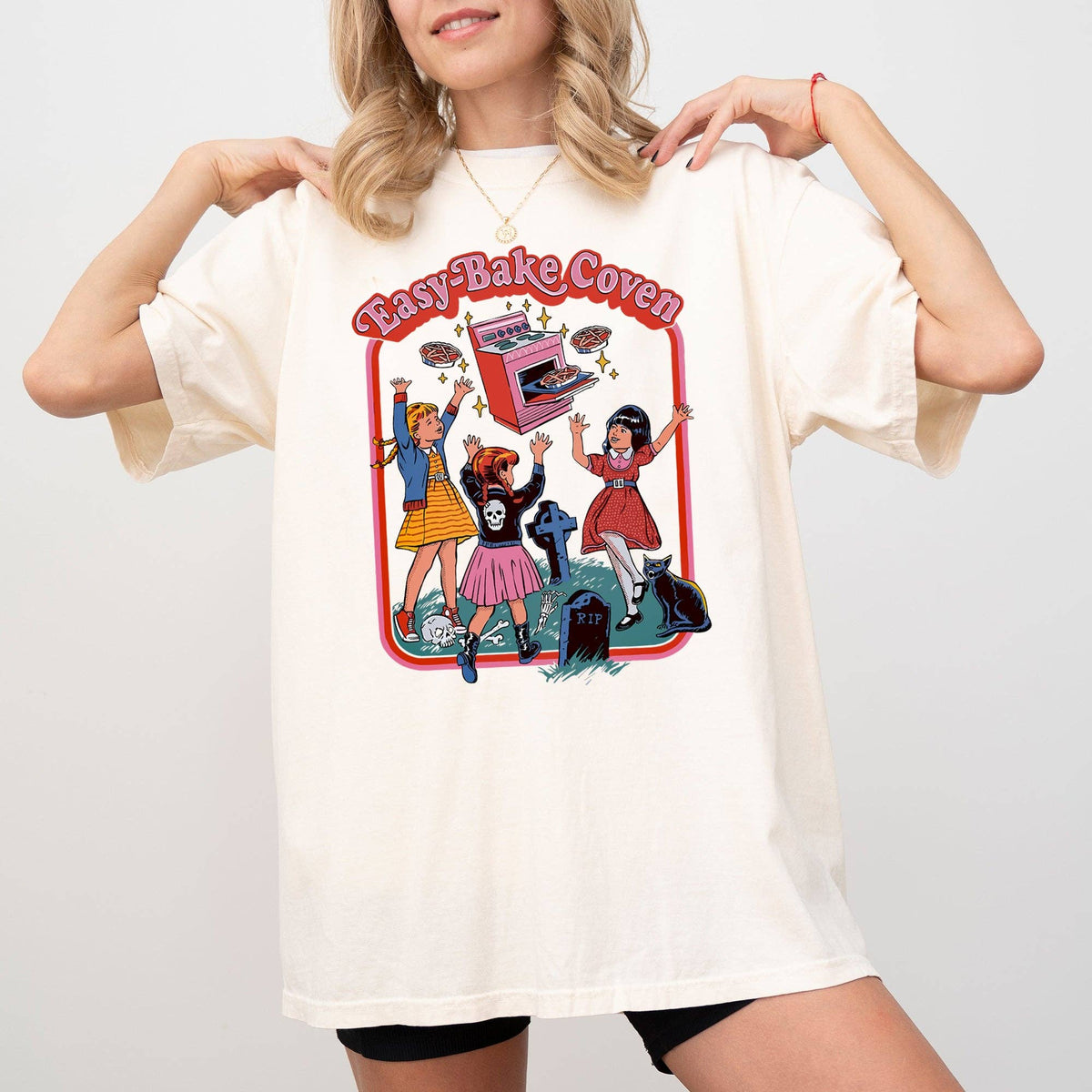 A woman wearing an oversized cream-colored t-shirt with a colorful retro graphic that reads 'Easy Bake Oven' and features cartoon girls baking with a pink oven. The background is a clean, neutral studio gray.