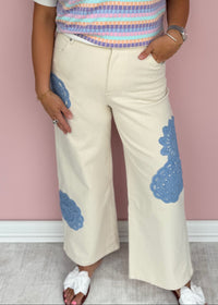 Woman wearing a colorful striped sweater and white pants with blue patterns against a pink wall.