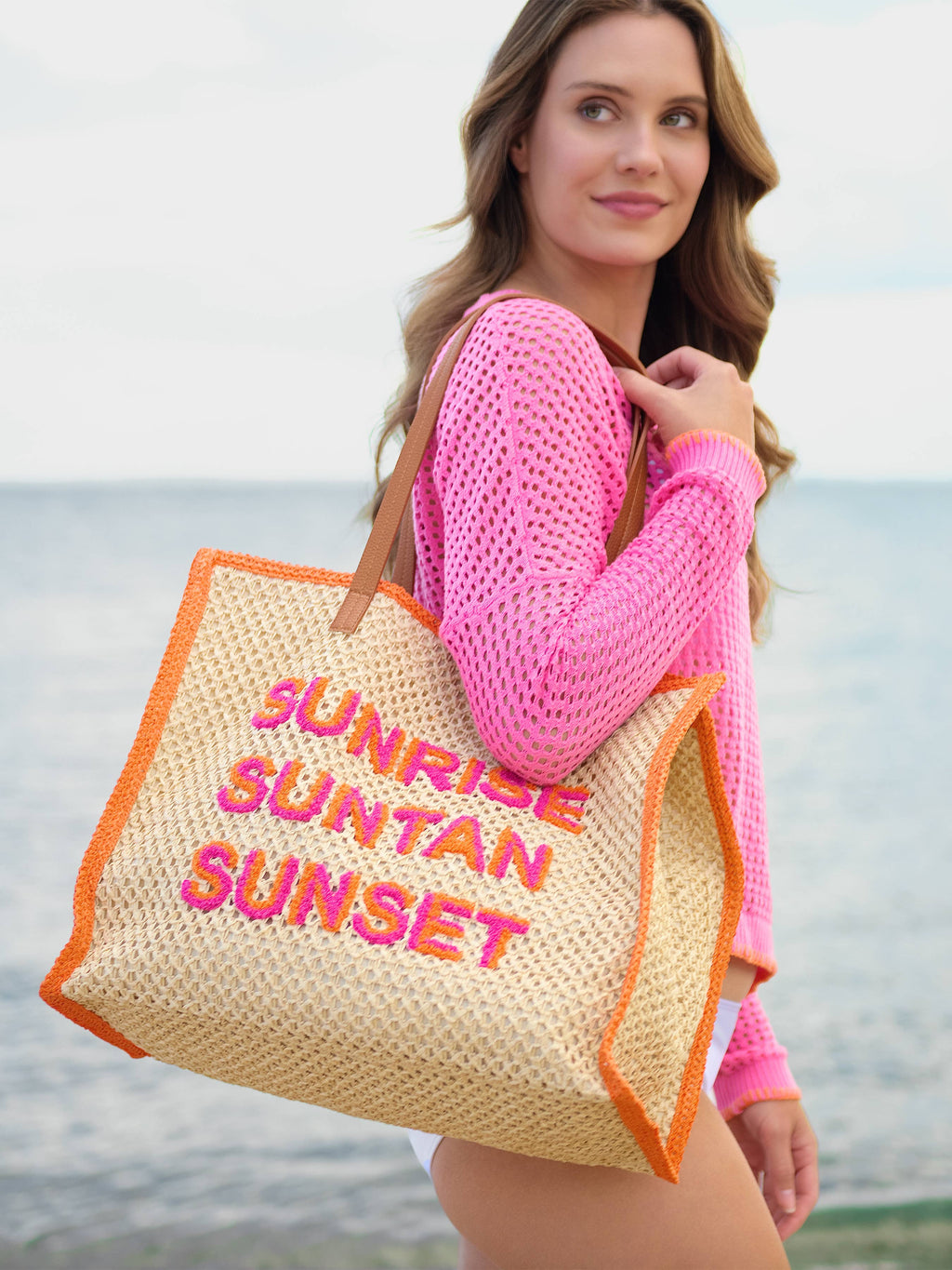 Person holding a straw bag with 'Sunrise Suntan Sunset' text on a beach.