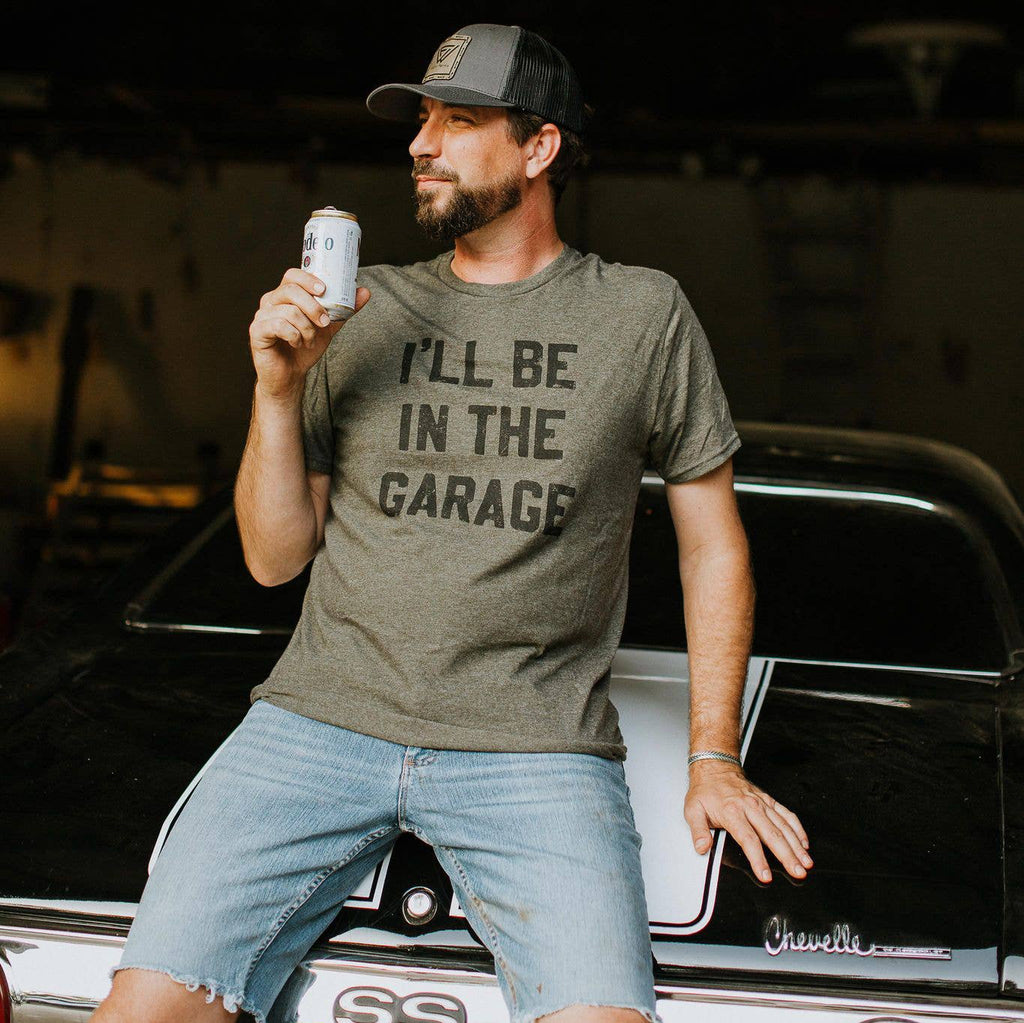 Man wearing a t-shirt with 'I'll be in the garage' text, holding a can, standing next to a car.