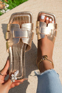 Gold metallic sandals with a textured finish worn by a person on a light background.