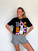 Person wearing a black t-shirt with colorful text and checkered shorts against a white brick wall.