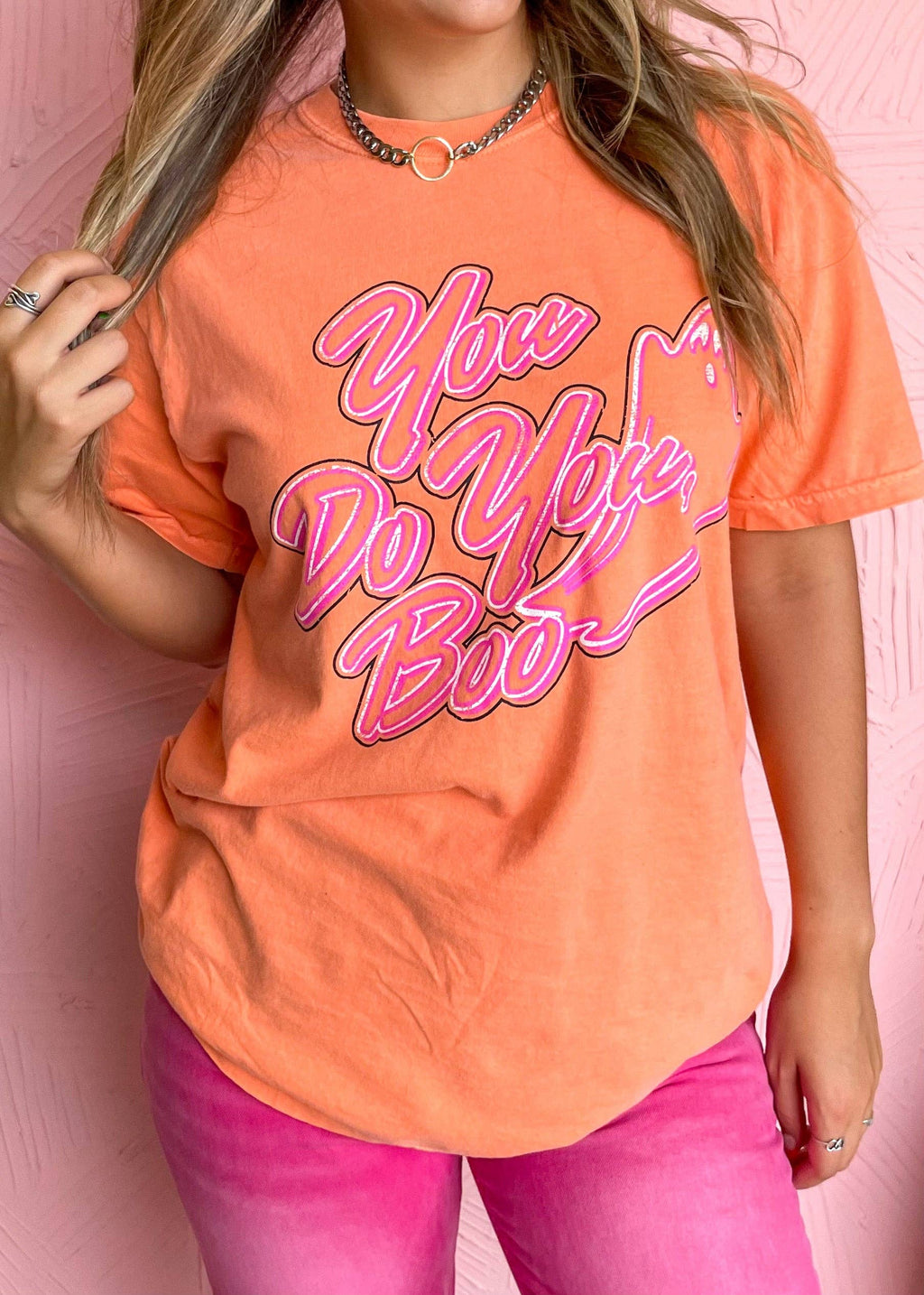 Person wearing an orange t-shirt with pink text and pink pants against a pink background