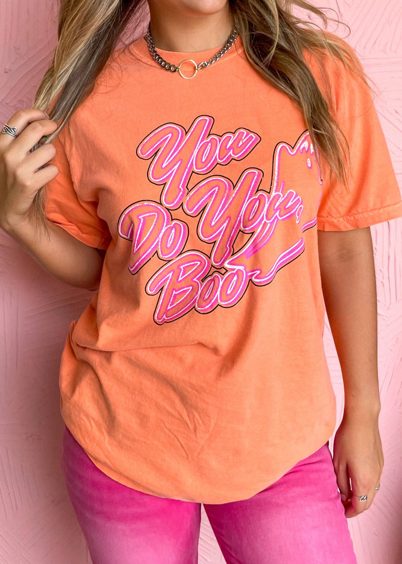 Person wearing an orange t-shirt with pink text and pink pants against a pink background