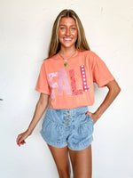 Woman wearing a coral t-shirt with 'FALL' printed on it and denim shorts against a white background