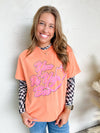 Woman wearing an orange shirt with pink text and checkered sleeves against a white textured wall.