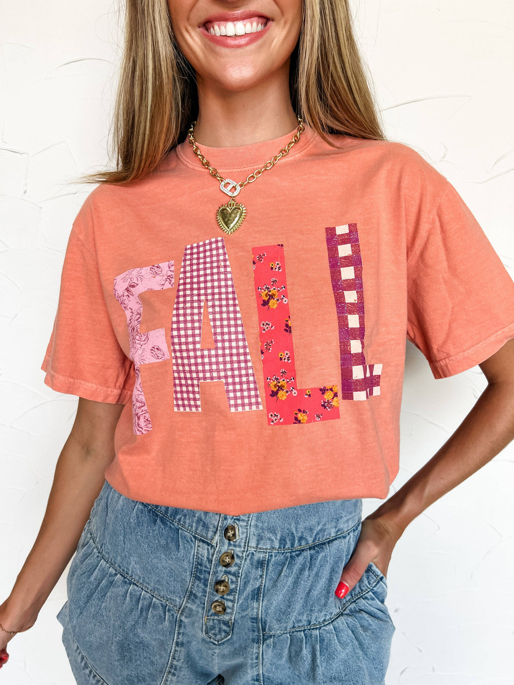 Person wearing a peach-colored t-shirt with 'FALL' text in checkered and floral patterns, paired with blue jeans.