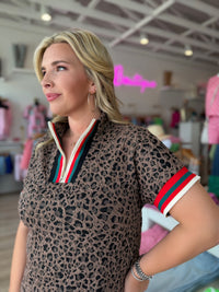 A woman wearing a short-sleeve leopard-print polo shirt with a green, white, and red striped collar and sleeve trim, standing in a boutique.