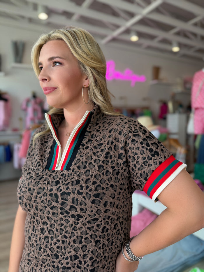 A woman wearing a short-sleeve leopard-print polo shirt with a green, white, and red striped collar and sleeve trim, standing in a boutique.