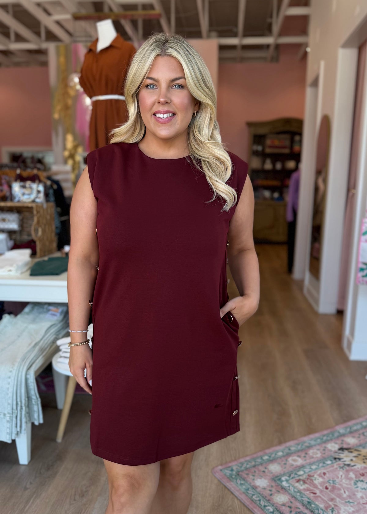 A woman modeling a sleeveless, crew-neck mini dress in deep burgundy with side pockets, standing inside a boutique with wood floors and pink walls.