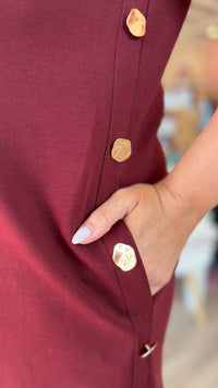 Close-up of a burgundy dress with large, textured gold buttons and a side pocket, shown on a model with a hand in the pocket.