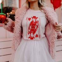 A person wearing a pink furry jacket and a pink skirt holding a green cup, with a red graphic of a queen on a playing card printed on a pink t-shirt underneath.
