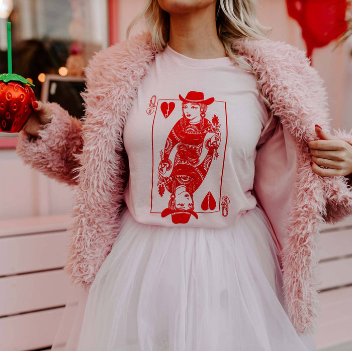 A person wearing a pink furry jacket and a pink skirt holding a green cup, with a red graphic of a queen on a playing card printed on a pink t-shirt underneath.