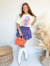 Woman wearing a colorful graphic t-shirt, purple skirt, white boots, and holding an orange handbag.
