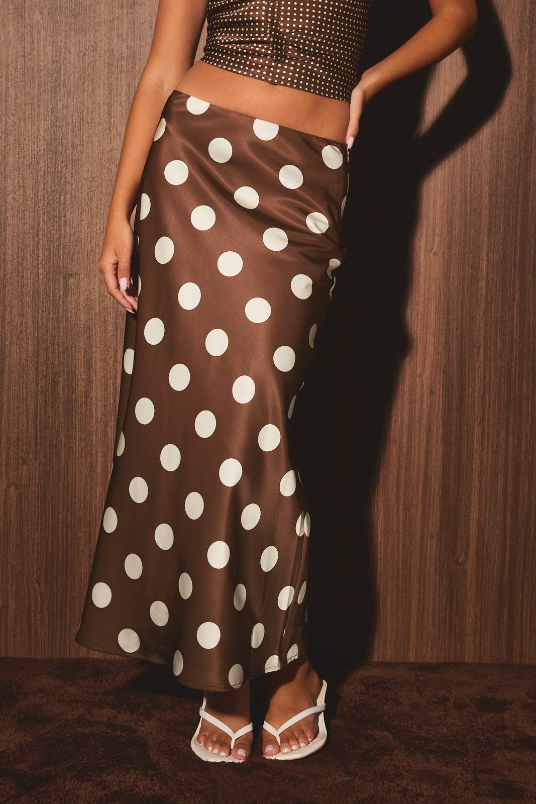 A woman wearing a brown satin maxi skirt with large white polka dots, paired with a matching brown studded crop top and white thong sandals, standing against a dark wood-paneled wall.