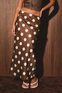 A woman wearing a brown satin maxi skirt with large white polka dots, paired with a matching brown studded crop top and white thong sandals, standing against a dark wood-paneled wall.